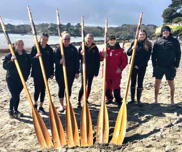 Ladies B crew stood in a line on the beach with their oars resting on their shoulders. 